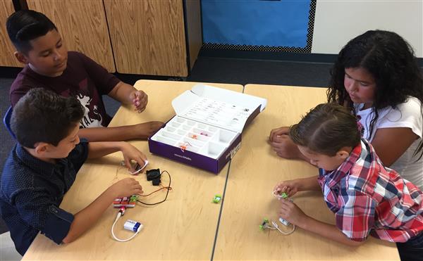 Four elementary students -- three boys and one girll -- work with electronic components, while sitting at two facing desks